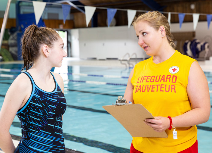 A lifeguard holding a clipboard speaks to a young female swimmer.