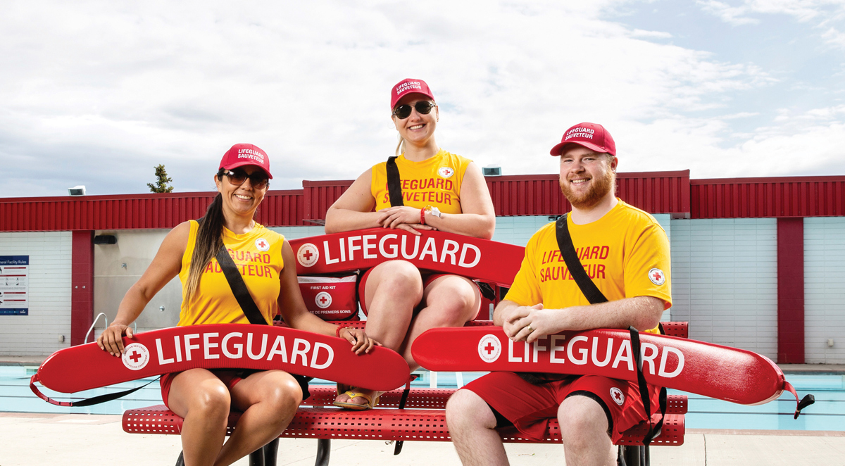 Three lifeguards pose with red rescue tubes in front of an outdoor pool.