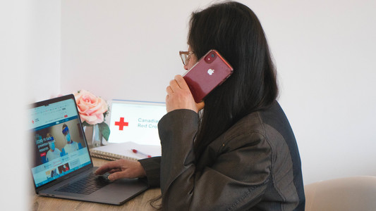 A woman sitting at a desk, using a mobile phone and also visiting the Red Cross homepage.