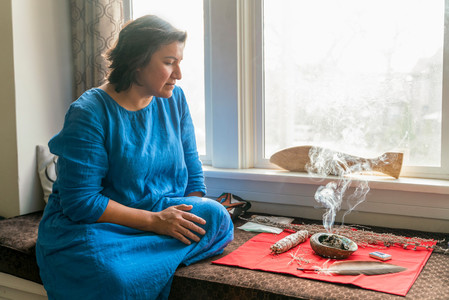 An indigenous woman sits by a window, burning sage.