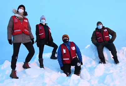 Four Canadian Red Cross volunteers wearing red vests over top of their winter coats climb a mountain of snow. All of them are wearing medical face masks.