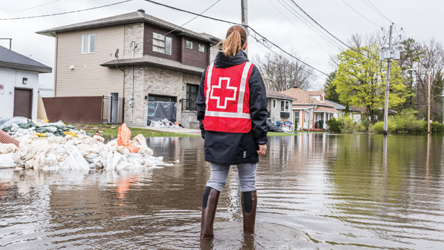 A Red Cross representative wearing a red vest surveys the flooding of a rural street in a neighborhood.