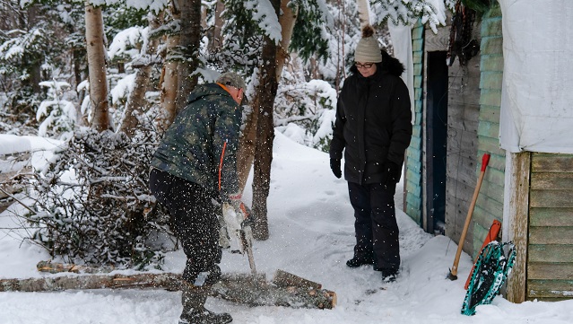 Outside of a shed in winter, an adult is chopping a log with an axe while wearing winter gear.