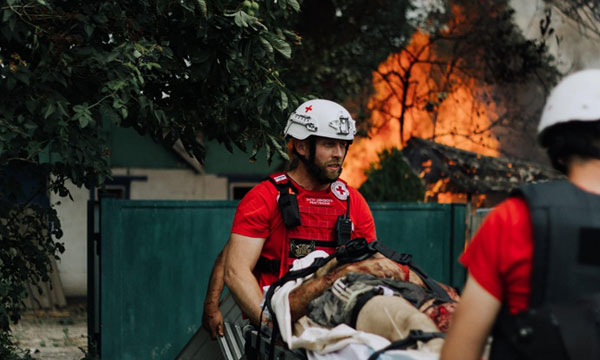 Two Red Cross medics carry an injured person on a stretcher, as a fire engulfs a tree and a building in the background.