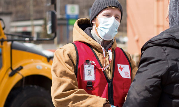 A Canadian Red Cross volunteer wears a medical mask, and is standing outside in a parking lot speaking to another person.