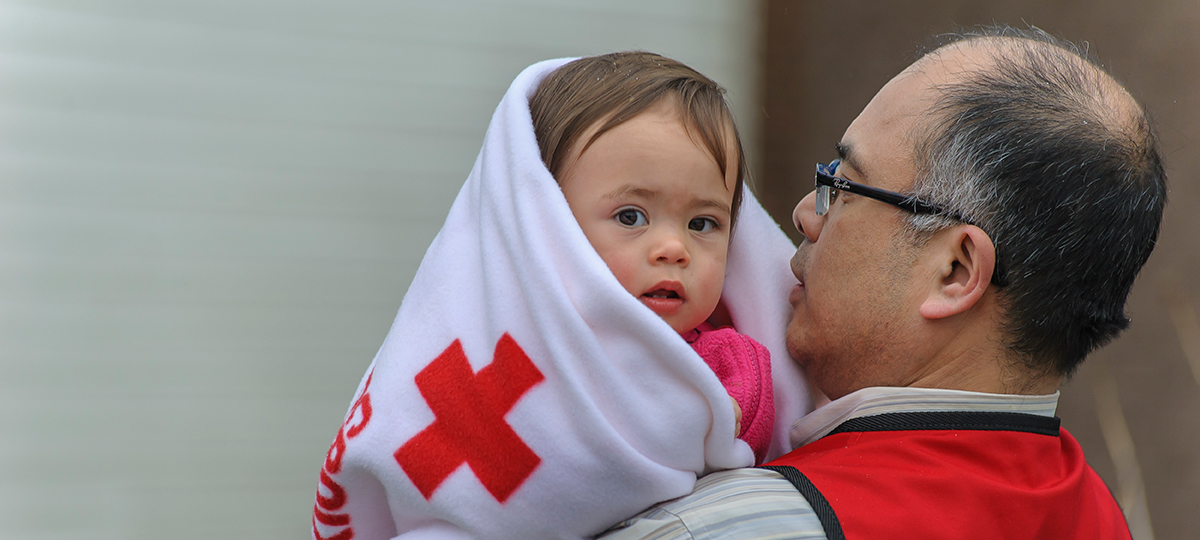 An older male Red Cross volunteer provides comfort to a toddler wrapped in a Red Cross fleece blanket.