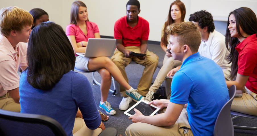 A group of high school students sitting in a circle, having a discussion and taking notes on electronic devices.