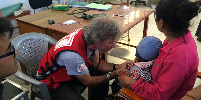 A Red Cross representative wearing a red vest uses a stethoscope on an infant, being held in their mother's lap.