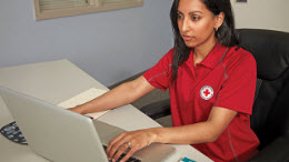 A woman wearing a red Canadian Red Cross polo sits at a desk, working on a laptop.