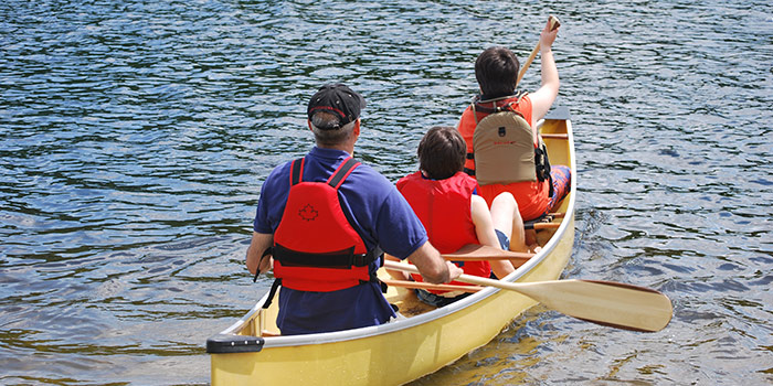 Two children and an adult man, all wearing life jackets, navigate a lake inside of a canoe.