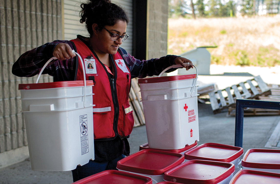 A young woman wearing a Red Cross vest lifts two large buckets of supplies.