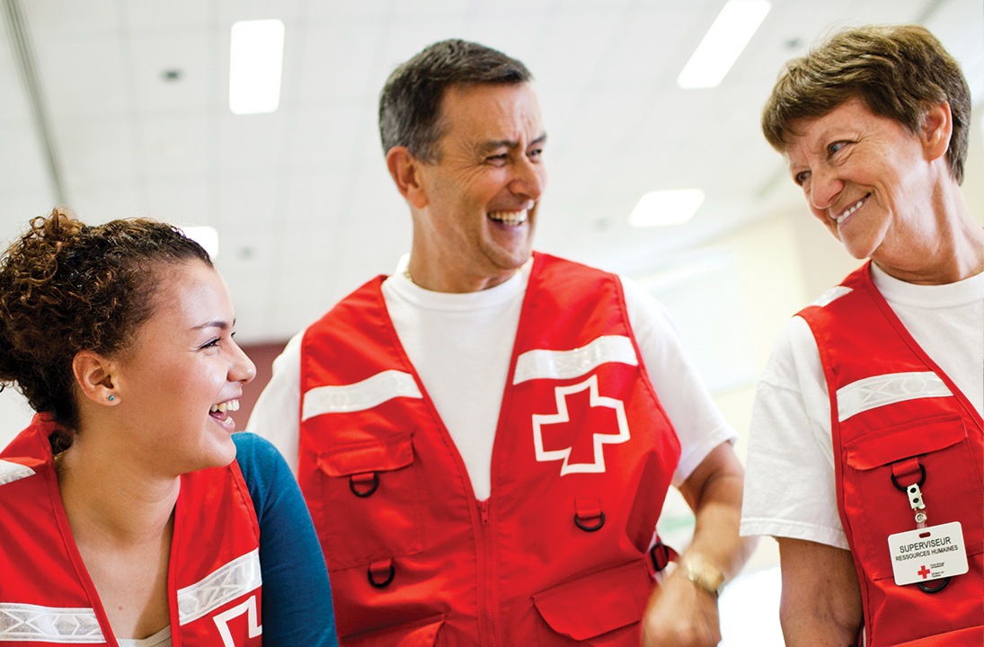 Three Red Cross volunteers wearing red vest smile at each other.