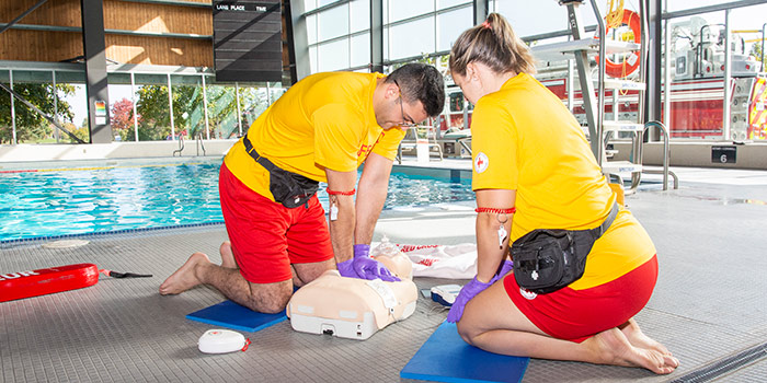 Two lifeguards inside an indoor pool demonstrate proper CPR technique on a dummy.