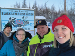 Four people in outdoor winter gear stand in front of Black Lake town sign in winter