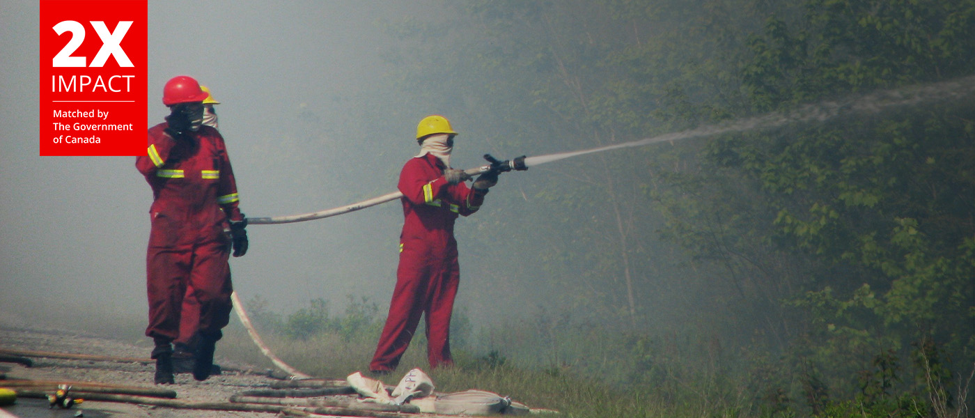 Firemen douse fires with water, with smoke in the background.