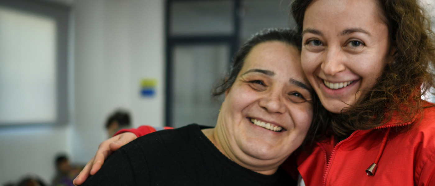 A smiling Red Cross volunteer offers a comforting embrace to a hopeful-looking woman.