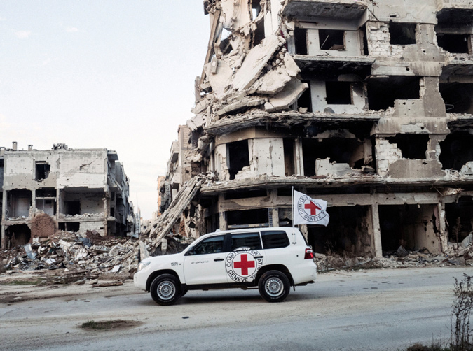 An SUV adorned with a Red Cross logo drives by the crumbling ruins of a building.