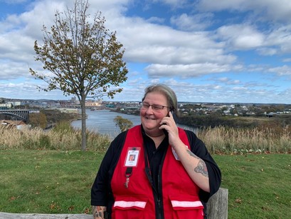 A woman in a Red Cross vest smiling outside, holding a phone to ear