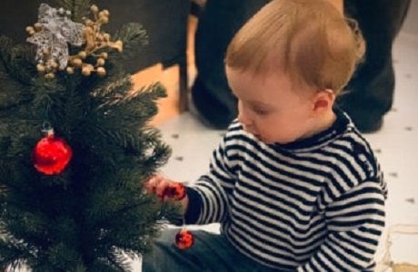 A baby sitting next to an artificial Christmas tree, touching one of the bulbs