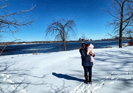 A woman holding a baby in winter wear standing on snowy ground in front of a body of water