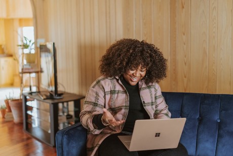 A woman with dark curly hair is sitting on a couch with a computer and seems to be talking to someone on facetime