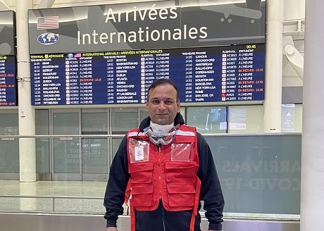 A man in a Canadian Red Cross vest smiling from inside an airport with a sign that says Arrivals.