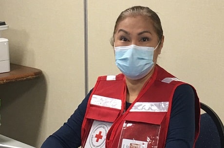 A woman in a Red Cross vest sitting at a computer desk, smiling behind a mask