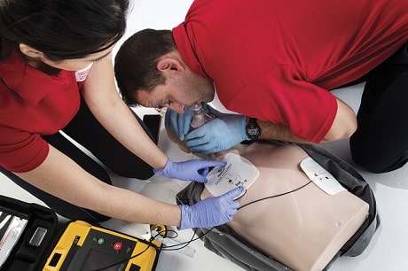 Two people in red shirts around a mannequin applying an AED to it