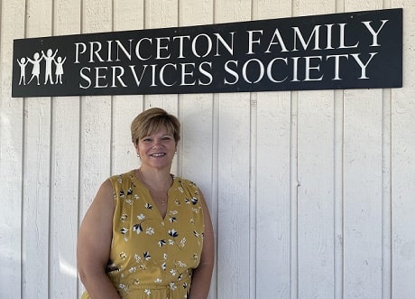 A woman in a yellow shirt smiling while leaning against a white wall with a sign that says: Princeton Family Services Society