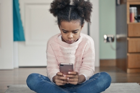 A girl sits on the floor in a home setting and looks at a mobile phone in her hands. Credit: bruce mars / unsplash.com 