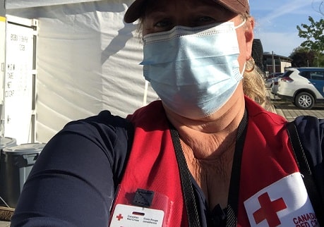 A selfie of a woman in a baseball cap, mask and Red Cross vest in front of a white tent.