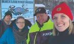 Four people in outdoor winter gear stand in front of Black Lake town sign in winter