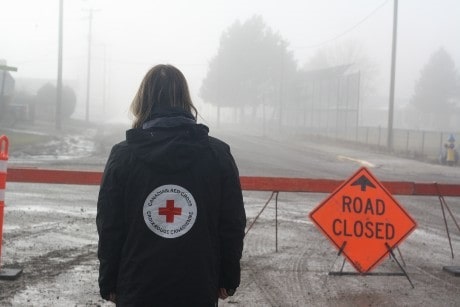 A person in a Red Cross jacket looking at a closed road due to flooding