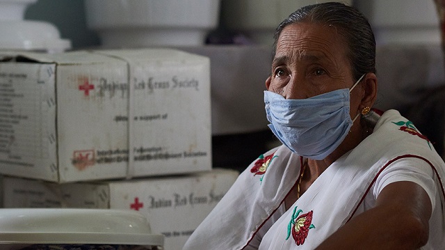 A woman wearing a medical mask sits surrounded by Indian Red Cross Society boxes.