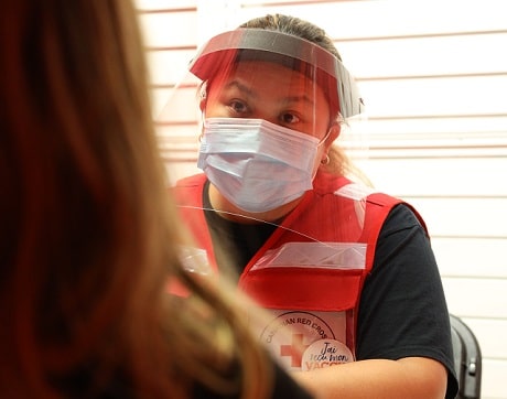 A woman in Red Cross vest wearing mask and face shield speaking to another woman