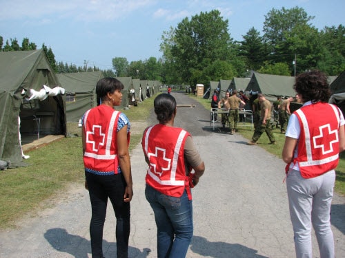 Three Red Cross volunteers standing outside, looking down a road of army encampment tents