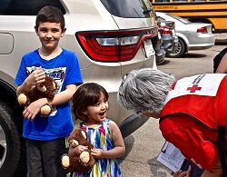 Red Cross volunteer talking to two young children, both holding teddy bears, outside in a parking lot
