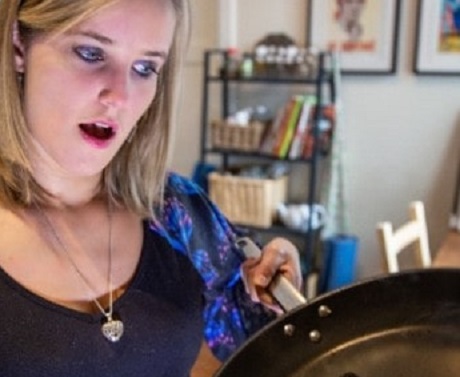 A woman looking shocked at burned food in a frying pan