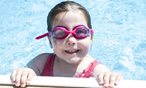 little girl with goggles in a pool holding onto the side and smiling at the camera
