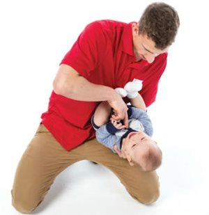 A man on his knees holds a baby on his back, using his free hand to deliver chest compressions using his fingers