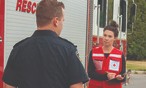 woman in vest with clipboard talks to male firefighter in front of a fire truck