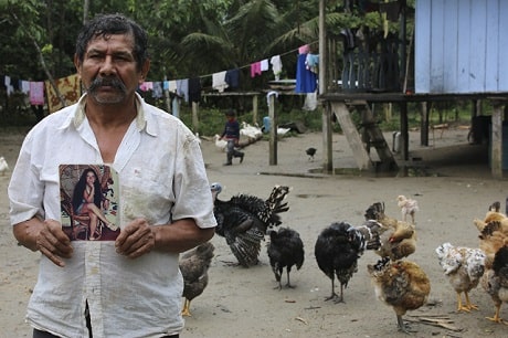 A man standing in a yard full of chickens, holding a photo of a young girl