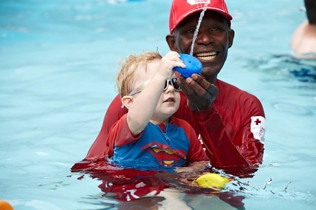 Barney holding a toddler in a swimming pool, smiling, while the child squirts water out of a toy.