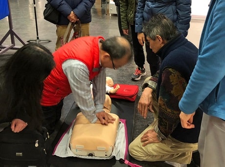 A man in a red vest is showing how CPR is done, on a mannequin, with a small crowd of people around him watching.