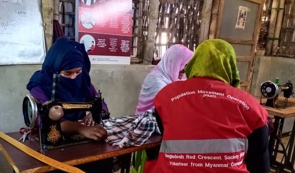 Two women work on sewing machines while a member of the Bangladesh Red Crescent talks to them.