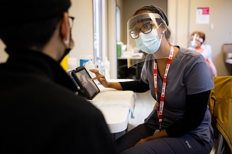 A person in face shield, mask, glasses and medical gown talking to another person
