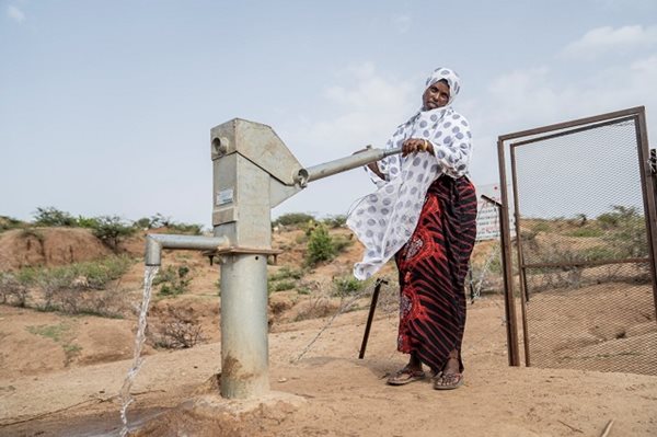 A woman standing near a water borehole in a barren field.