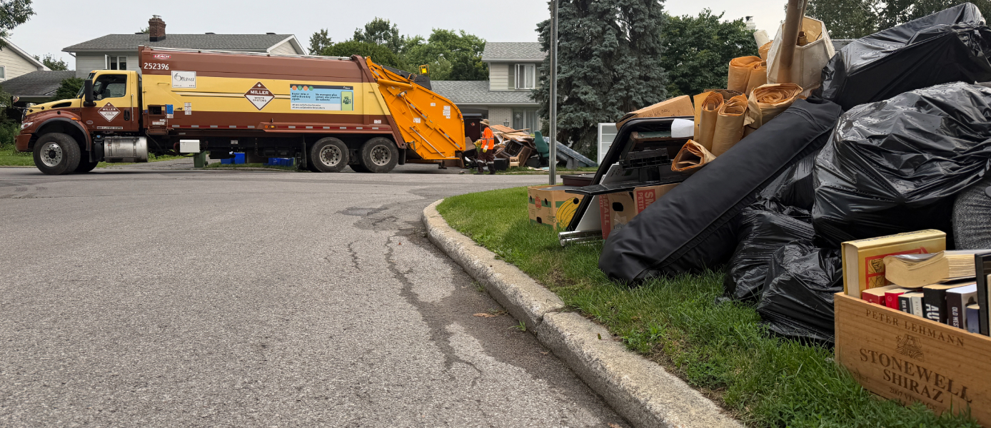 Garbage bags and other materials sit in front of homes with a garbage truck in the background.