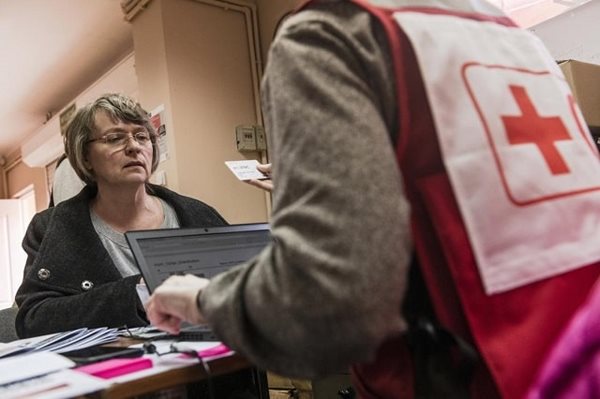A person in a Red Cross vest handing inputting information into a laptop provided by an older woman