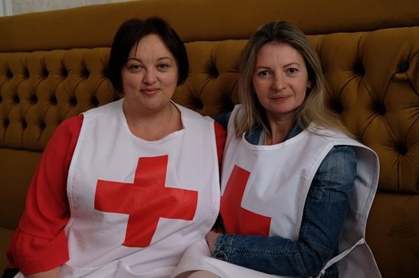 Two women in Red Cross vests sitting together on a large brown couch
